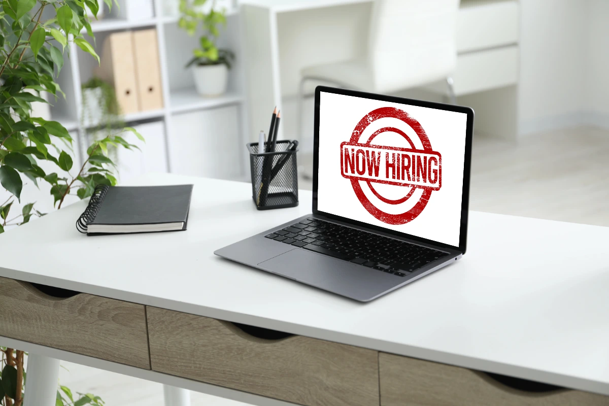 A laptop on a clean desk displaying a Now Hiring graphic, representing the concept of your website working as an employee for your business